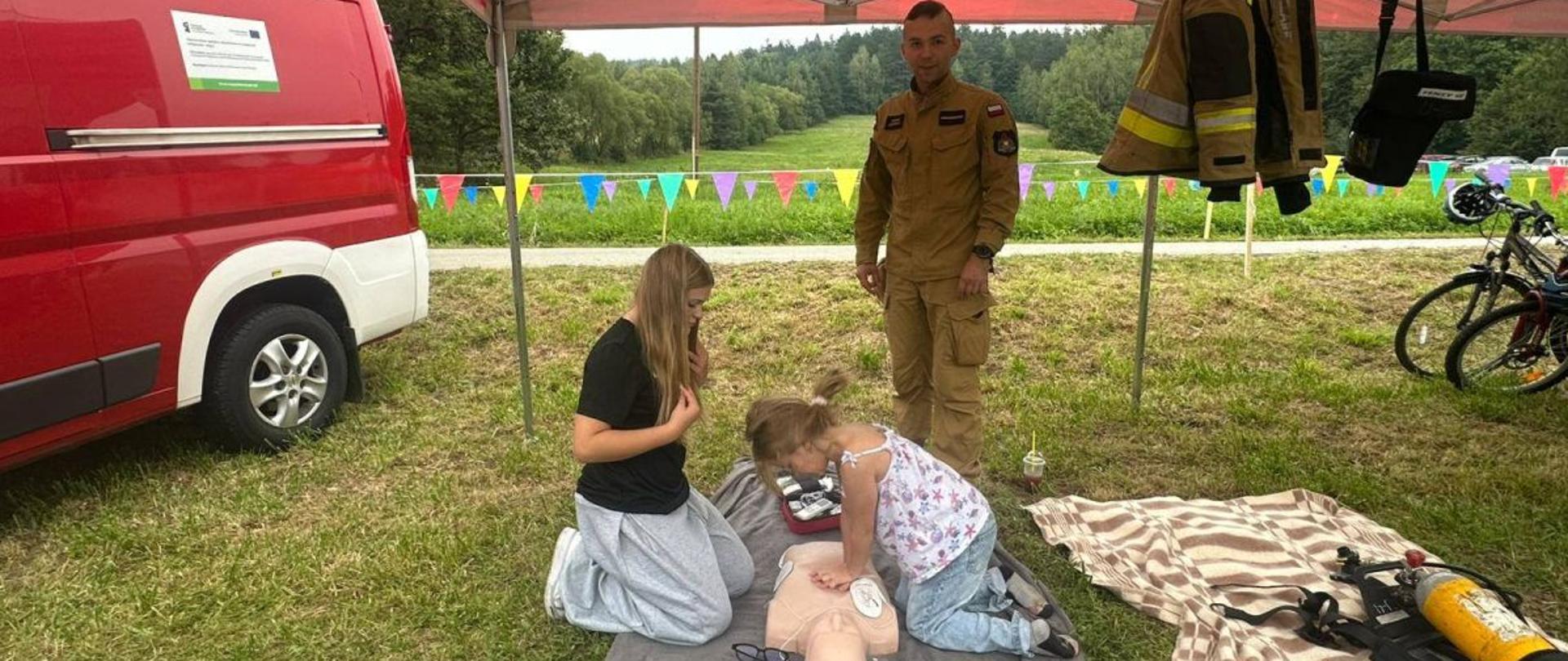 Zdjęcie przedstawia strażaka PSP i dwie dziewczynki pod namiotem stelażowym. Dziewczynki klęczą nad manekinem a strażak stoi. Jedna z dziewczyn uciska klatkę piersiową manekina. Pod namiotem znajduje się także torba medyczna, aed treningowe oraz ubranie strażackie specjalne. Obok namiotu stoi bus. 