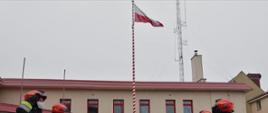 Strażacy stoją w dwóch rzędach na przeciwko siebie, uroczyście podnosząc flagę Rzeczypospolitej Polskiej na maszt.