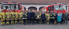 Wspólne zdjęcie funkcjonariuszy oraz pracowników cywilnych komendy na tle pojazdów pożarniczych i budynku komendy.