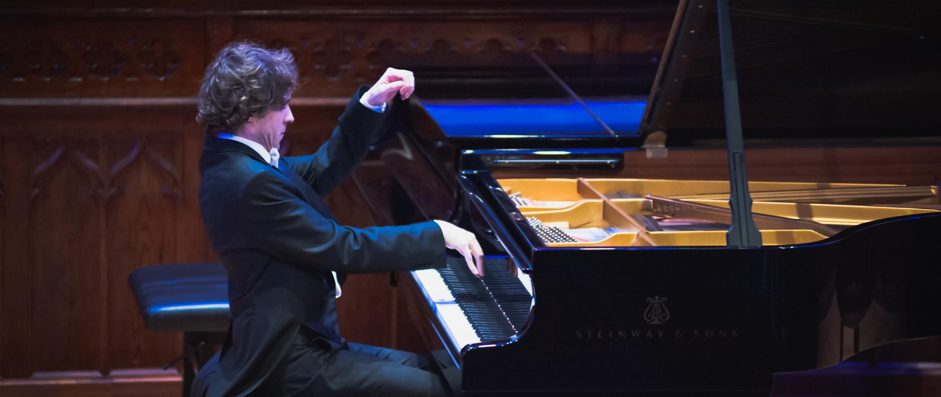 zdjęcie przedstawia pianistę Rafała Blechacza przy fortepianie na Sali królewskiej Państwowej Szkoły Muzycznej w Mielcu, w niebieskawych odcieniach oświetlenia scenicznego