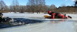 Dwóch strażaków w skafandrach wypornościowych podczas ćwiczeń na lodzie. Strażak po lewej w czarnym skafandrze znajduje się w przeręblu, strażak po prawej w czerwonym skafandrze w pozycji leżącej przesuwa się do tego pierwszego. 