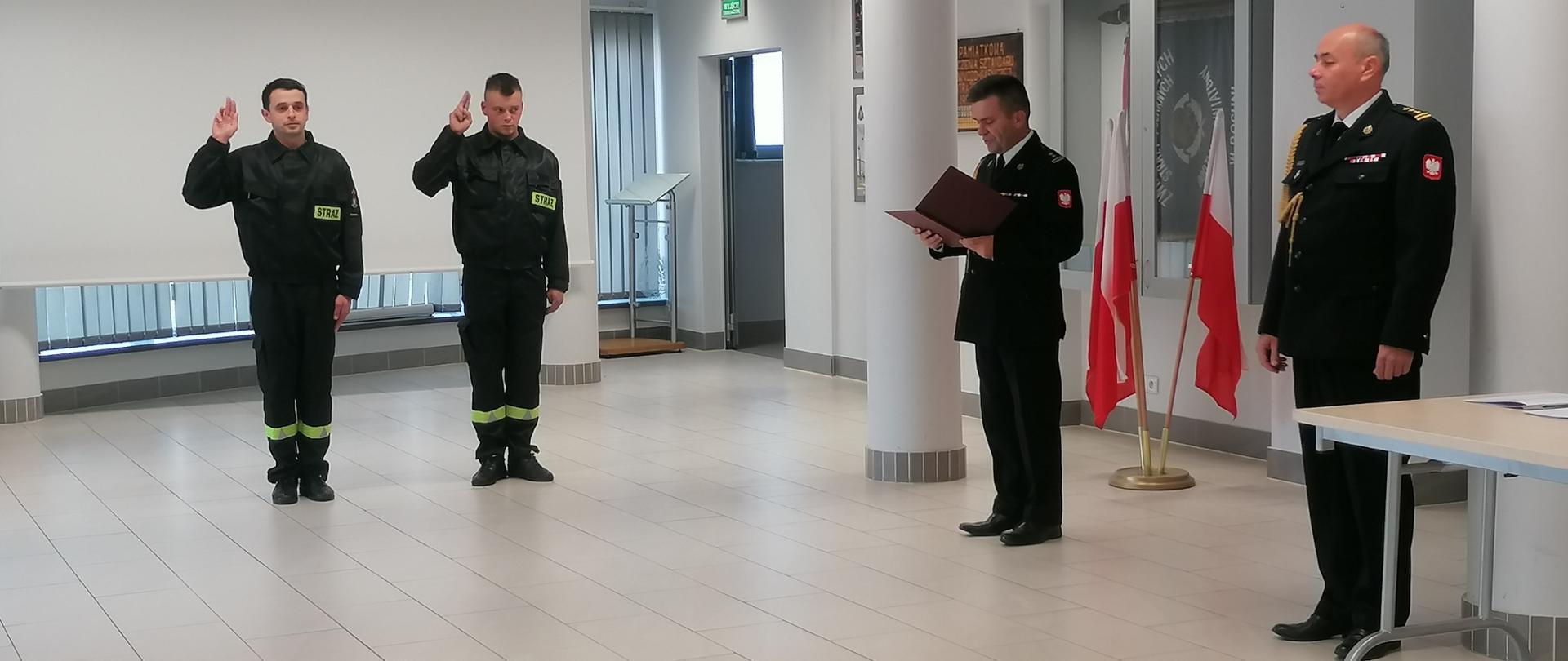 Na zdjęciu widzimy strażaków podczas uroczystej zbiórki z okazji ślubowania nowo przyjętych do służby strażaków. Dwóch strażaków składających ślubowanie ma uniesioną prawą rękę z dłonią na wysokości oczu z palcami ułożonymi jak do salutowania. Z prawej strony zdjęcia stoi Komendant Powiatowy Państwowej Straży Pożarnej w Bochni odczytujący rotę ślubowania, obok niego stoi Zastępca Komendanta Powiatowego Państwowej Straży Pożarnej w Bochni. Komendanci występują w mundurach wyjściowych, a strażacy składający ślubowanie w ubraniach koszarowych.
