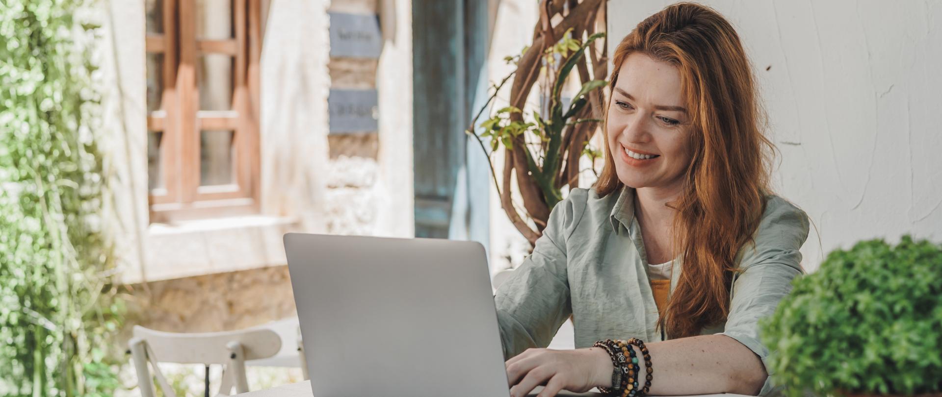 A woman working on a laptop at an outdoor table, surrounded by greenery. She is focused and smiling, enjoying remote work in a relaxed setting.