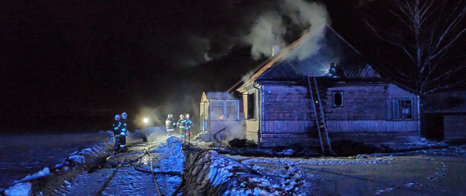 Wieczorem na zaśnieżonym podwórku stoją strażacy z rozwiniętymi wężami. Po prawej stronie budynek mieszkalny z którego wydobywa się dym.