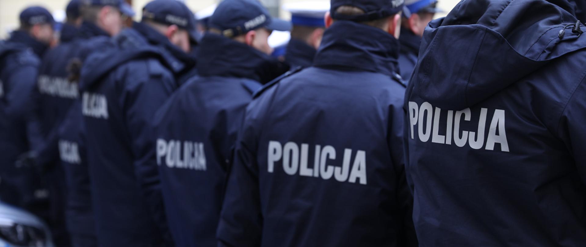 Na zdjęciu widać stojących w rzędzie policjantów w kurtkach z widocznym na plecach napisem "POLICJA". 