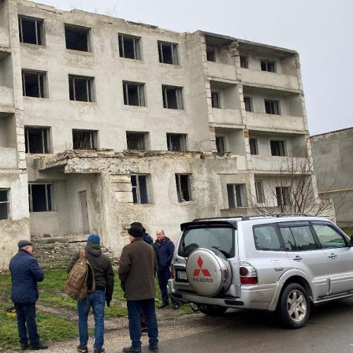 Kilka osób stoi przed bardzo zaniedbanym blokiem mieszkalnym, obok zaparkowanych dwóch samochodów.