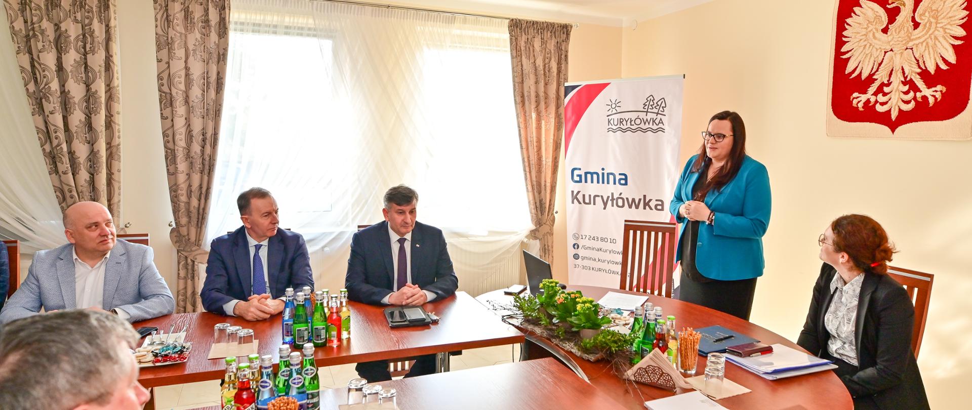 Grupa osób w sali siedzi przy stołach. Obok stoi wiceminister Małgorzata Jarosińska-Jedynak.