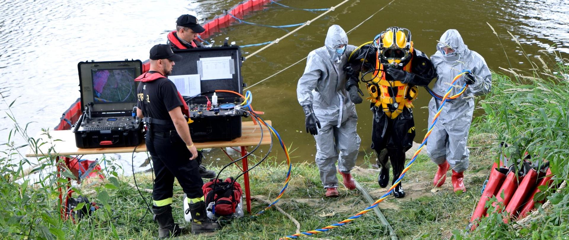 Zdjęcie zrobione na zewnątrz. Dwóch strażaków ubranych stroje ochrony przeciwchemicznej pomaga strażakowi płetwonurkowie wyjść na brzeg rzeki. Obok widać dwóch strażaków obsługujących sprzęt niezbędne do komunikacji z płetwonurkiem.