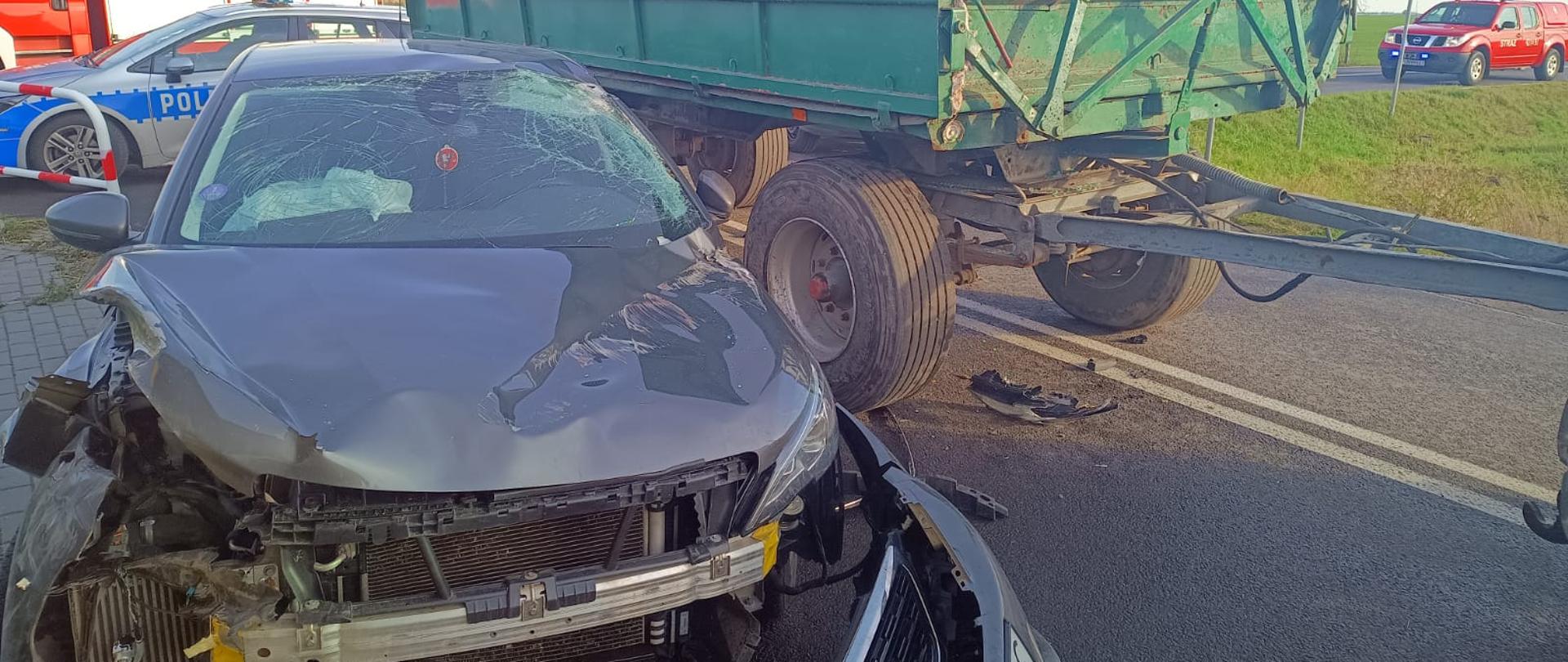 Na zdjęciu widać uszkodzony przód samochodu osobowego. Z prawej strony zdjęcia, obok auta znajduje się przyczepa rolnicza. W tle widać dwa samochody pożarnicze oraz radiowóz policyjny