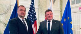 Wiceminister Tomasz Rzymkowski podaje rękę mężczyźnie ubranemu w garnitur. W tle flagi amerykańskie i Unii Europejskiej. 