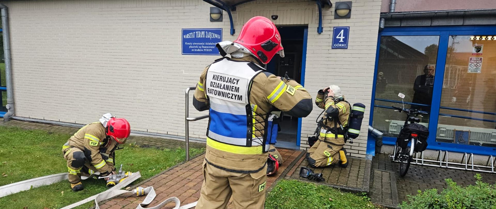 Strażacy ubierają sprzęt ochrony układu oddechowego przed wejściem do budynku.