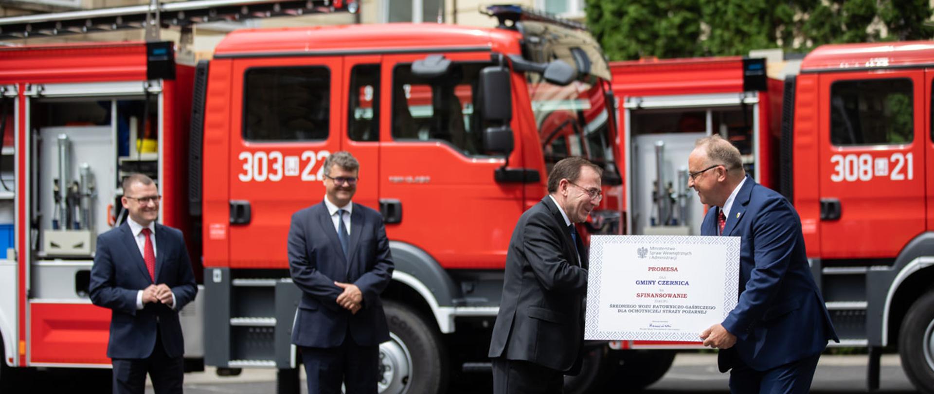 Na zdjęciu widać jak Mariusz Kamiński minister SWiA wręcza jedną z promes przedstawicielowi Gminy Czernica. W tle widać również Macieja Wąsika i Pawła Szefernakera wiceministrów SWiA i dwa wozy strażackie stanowiące tło zdjęcia. 
