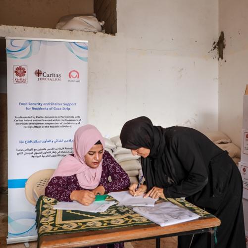 Jedna kobieta z głową zakrytą różową chustą siedzi przy stole, druga kobieta również z głową zakrytą czarną chustą pochyla się nad stołem i pisze na kartce papieru. W tle jest plakat z logo Polskiej pomocy i Caritas oraz białe pudełka. 