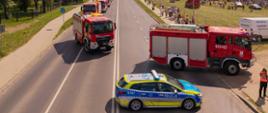 Kolumna wozów strażackich jadąca dwupasmową ulicą podczas festynu. Na pierwszym planie radiowóz policyjny blokujący ruch oraz duży wóz strażacki skręcający na pobliski teren zielony, gdzie zgromadzili się ludzie.
