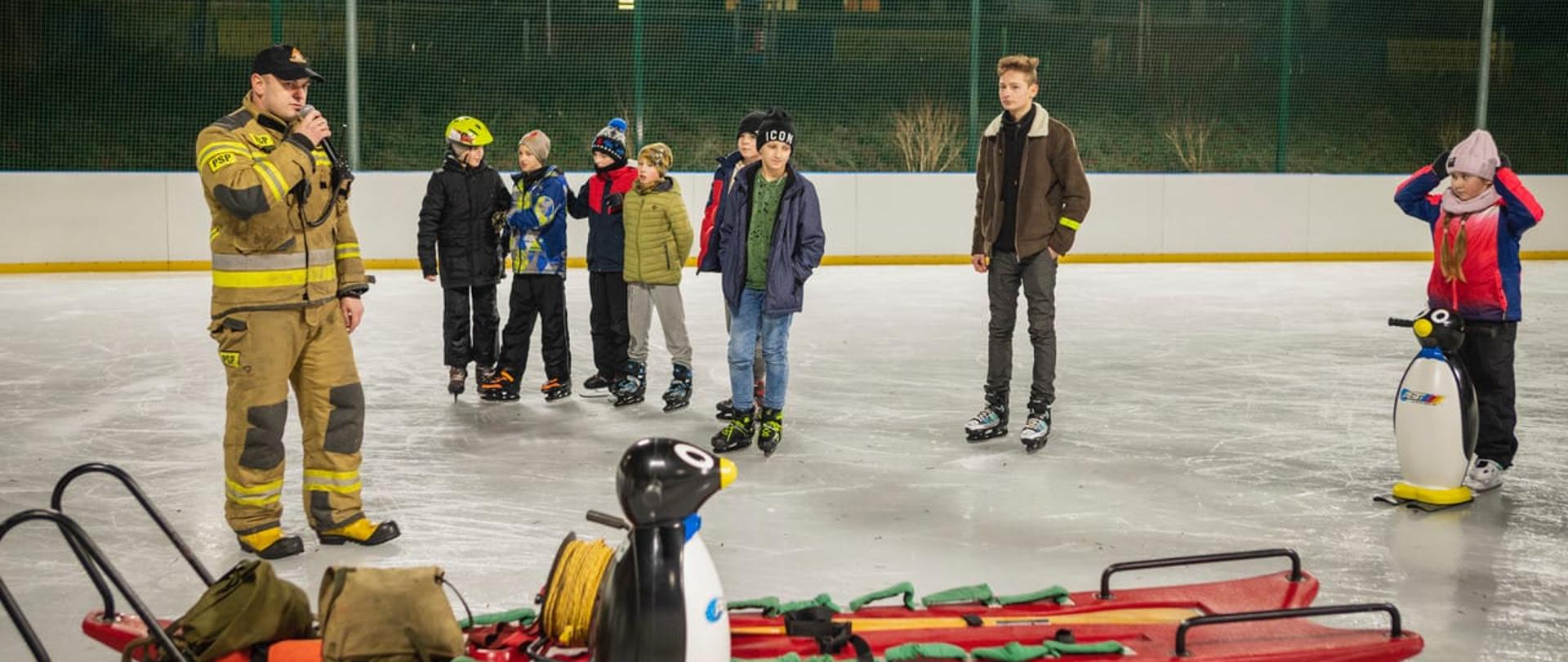 Zdjęcie przedstawia strażaka na tafli lodowej w tle widać dzieci na łyżwach oraz deskę lodową.