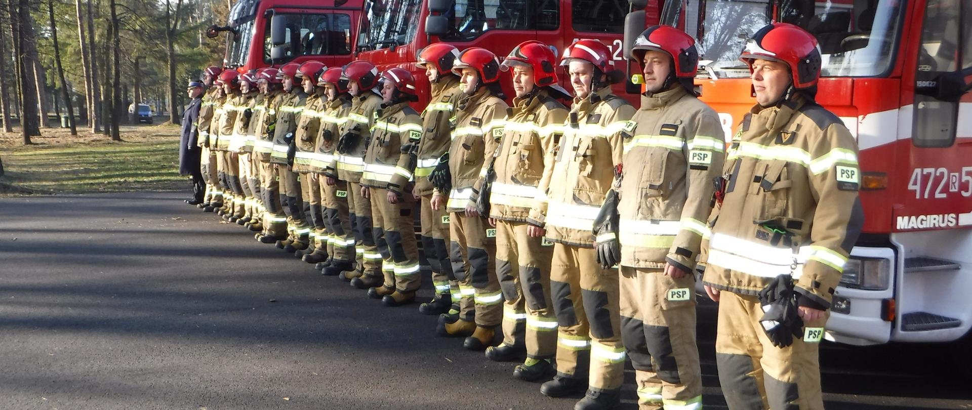Na zdjęciu widzimy strażaków z Komendy Powiatowej Państwowej Straży Pożarnej w Leżajsku, którzy w milczeniu stoją na baczność w szeregu na tle pojazdów pożarniczych. W ten sposób, uczcili minutą ciszy bohaterskich ratowników Ukrainy, którzy oddali swoje życie podczas służby w trudnych warunkach i dramatycznych dniach wojny.