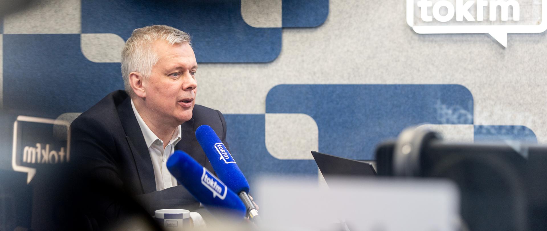 Na zdjęciu wnętrze studia radiowego. Po prawej stronie zdjęcia, siedzący przy stole minister Tomasz Siemoniak. Na stole znajdują się dwa niebieskie mikrofony z napisem "tokfm", kubek oraz laptop. Po lewej stronie zdjęcia logo radia "tokfm". Na górze zdjęcia biała kamera. Wnętrze w szaro-niebieskich prostokątach.