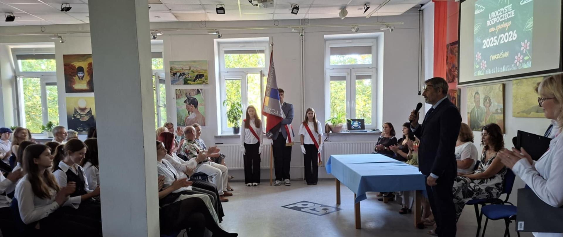 Zdjęcie przedstawia uroczystość inauguracji roku szkolnego 2025/2026, na pierwszym planie widać poczet sztandarowy po lewej stronie siedzący nauczyciele i uczniowie, po prawej dyrekcja szkoły