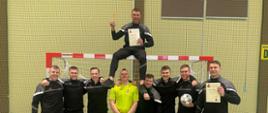 Na zdjęciu widoczna jest drużyna strażaków z Komendy Powiatowej PSP w Olecku, która zajęła IV miejsce podczas XX Mistrzostw Województwa Warmińsko-Mazurskiego Strażaków PSP w Futsalu. Zawodnicy ubrani są w czarne dresy z szarymi akcentami, stoją na tle bramki w hali sportowej.
