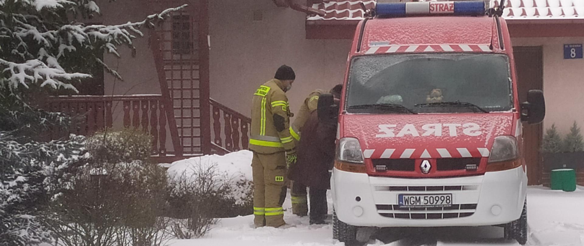 Dwóch strażaków pomaga starszej kobiecie wsiąść do lekkiego samochodu ratownictwa technicznego typu bus w kolorze czerwonym z białym zderzakiem należącego do OSP Żelechów, w tle widoczny budynek jednorodzinny. 