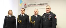 Funkcjonariusze Państwowej Straży Pożarnej. 3 mężczyzn oraz 1 kobieta. w umundurowaniach wyjściowych granatowych. na drugim planie logo Państwowej Straży Pożarnej