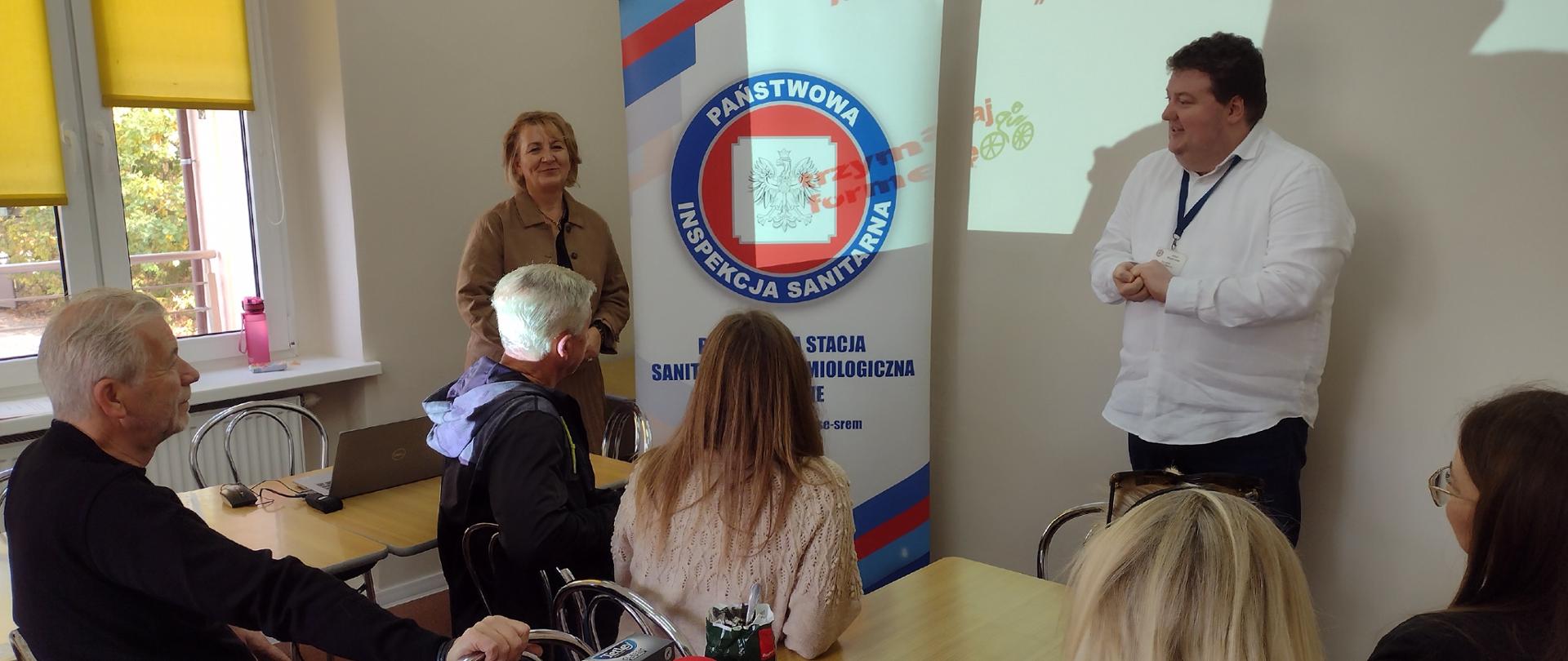 sala wykładowa, pięć osób siedzących przy stolikach, dwie osoby stojące, w tle baner Państwowej Inspekcji Sanitarnej w Śremie