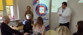 sala wykładowa, pięć osób siedzących przy stolikach, dwie osoby stojące, w tle baner Państwowej Inspekcji Sanitarnej w Śremie