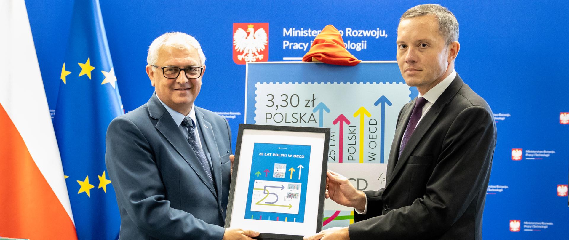 Grzegorz Piechowiak i Tomasz Zdzikot trzymają ramkę z pamiątkowym znaczkiem i kopertą.