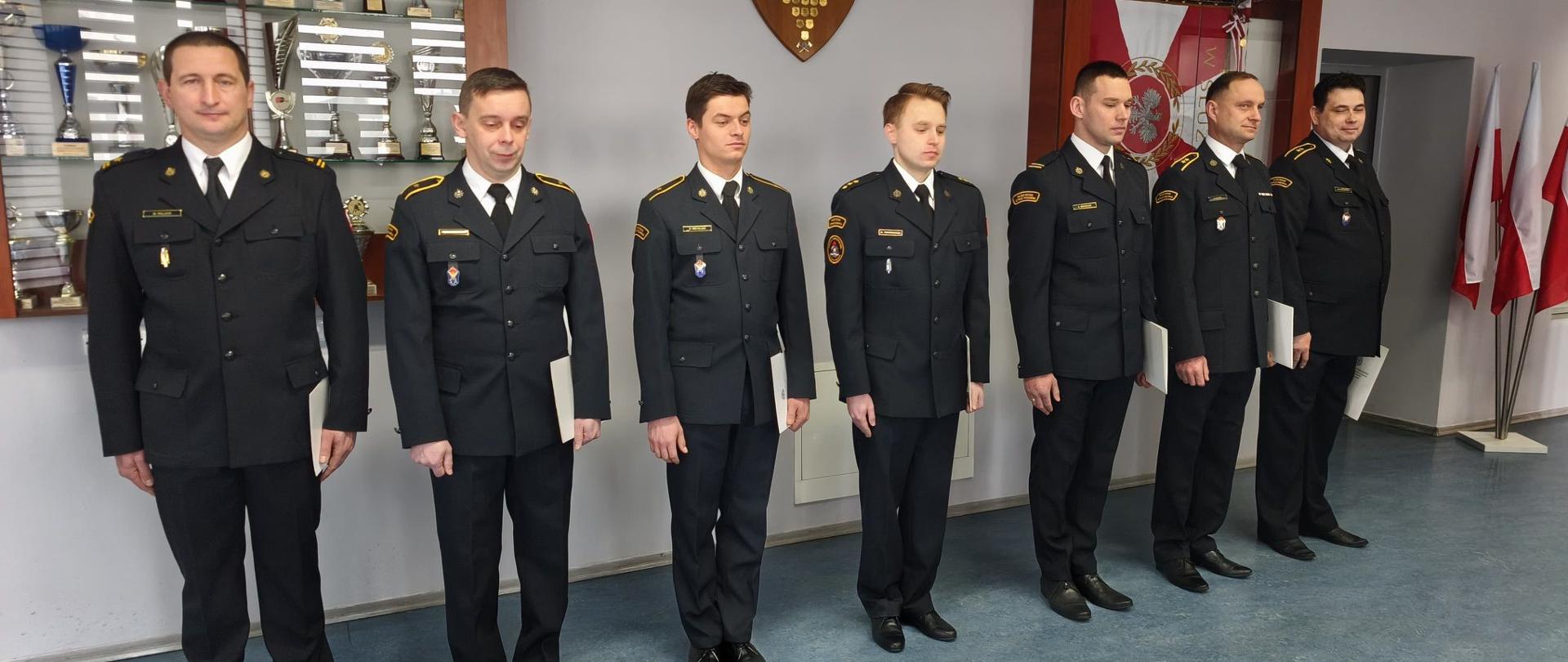 Na zdjęciu przedstawiono funkcjonariuszy Państwowej Straży Pożarnej ustawionych w jednym rzędzie w postawie zasadniczej. Każdy z nich trzyma dokument potwierdzający powierzenie nowego stanowiska lub awansu, będący wyrazem uznania za dotychczasową służbę, zaangażowanie oraz profesjonalizm