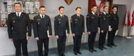 Na zdjęciu przedstawiono funkcjonariuszy Państwowej Straży Pożarnej ustawionych w jednym rzędzie w postawie zasadniczej. Każdy z nich trzyma dokument potwierdzający powierzenie nowego stanowiska lub awansu, będący wyrazem uznania za dotychczasową służbę, zaangażowanie oraz profesjonalizm