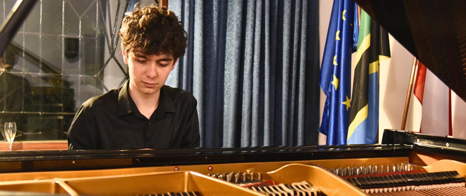 Mateusz Dubiel podczas europejskiego recitalu fortepianowego grający na fortepianie, z tylu flagi Unii Europejskiej, Tanzanii oraz Polski
