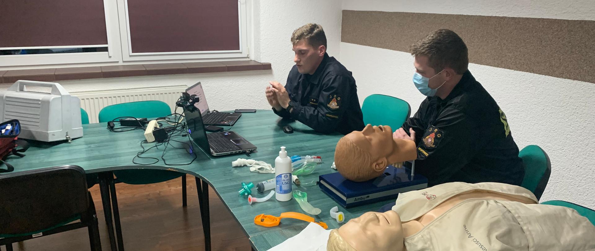 Na zdjęciu znajduje się dwóch strażaków w strojach koszarowych prowadzących wideokonferencje szkoleniową dla strażaków OSP KSRG. Zdjęcie wykonane od przodu prowadzących od ich lewej strony. Przed nimi na stole znajdują się dwa laptopy, a obok fantomy do resuscytacji i do nauki udrażniania dróg oddechowych. Przed strażakiem znajdującym się po prawej stronie na stole leżą rurki i maski krtaniowe do pokazów udrożniania dróg oddechowych. W tle okna świetlicy na której odbywa się wydarzenie.