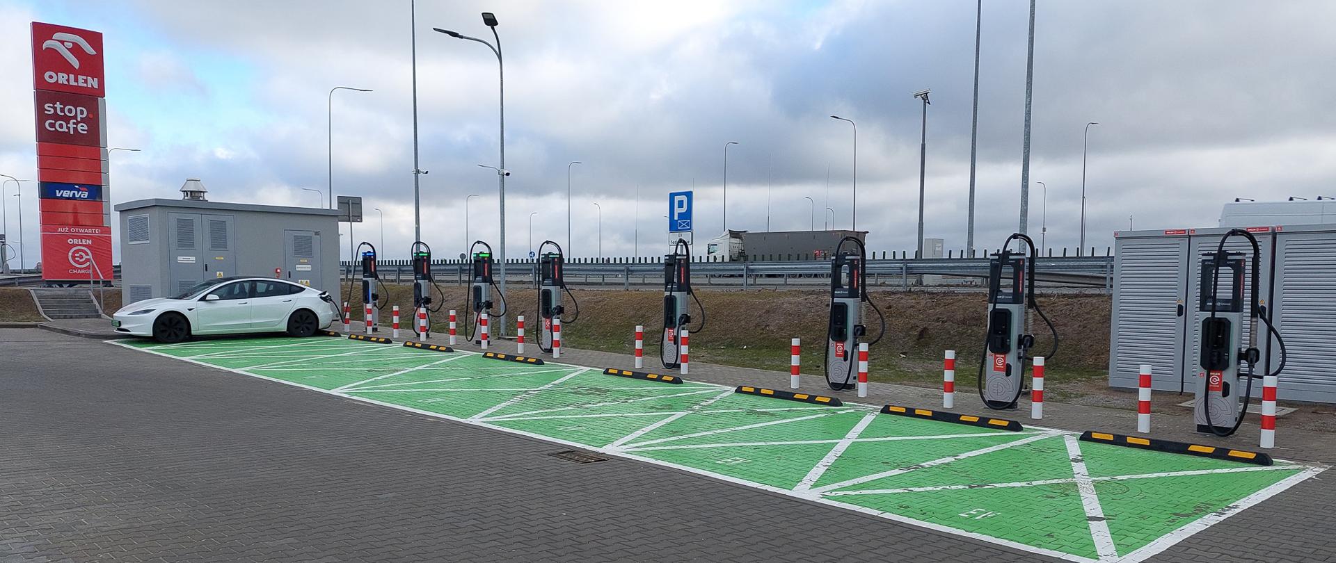 Strefa ładowania samochodów elektrycznych przy stacji Orlen z ośmioma stanowiskami szybkiego ładowania; na jednym z nich stoi biały samochód elektryczny. Zielone miejsca parkingowe oznaczone są jako przeznaczone do ładowania. W tle widoczne są lampy uliczne, drogowskazy i zabudowania techniczne.