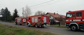 Na zdjęciu samochody ratowniczo-gaśnicze na drodze publicznej. Pojazdy w kolorze czerwonym. Posiadają sygnalizację ostrzegawczą. W dalszej części zabudowania gospodarcze. Zdjęcie wykonane w ciągu dnia. Zachmurzenie całkowite.