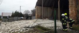 Na zdjęciu zawalona jedna ściana stodoły. Stodoła murowana c cegły kryta blachą. Strażacy w ubraniach specjalnych i hełmach zabezpieczają teren z boku widoczne ogrodzenie z siatki oraz płot betonowy.