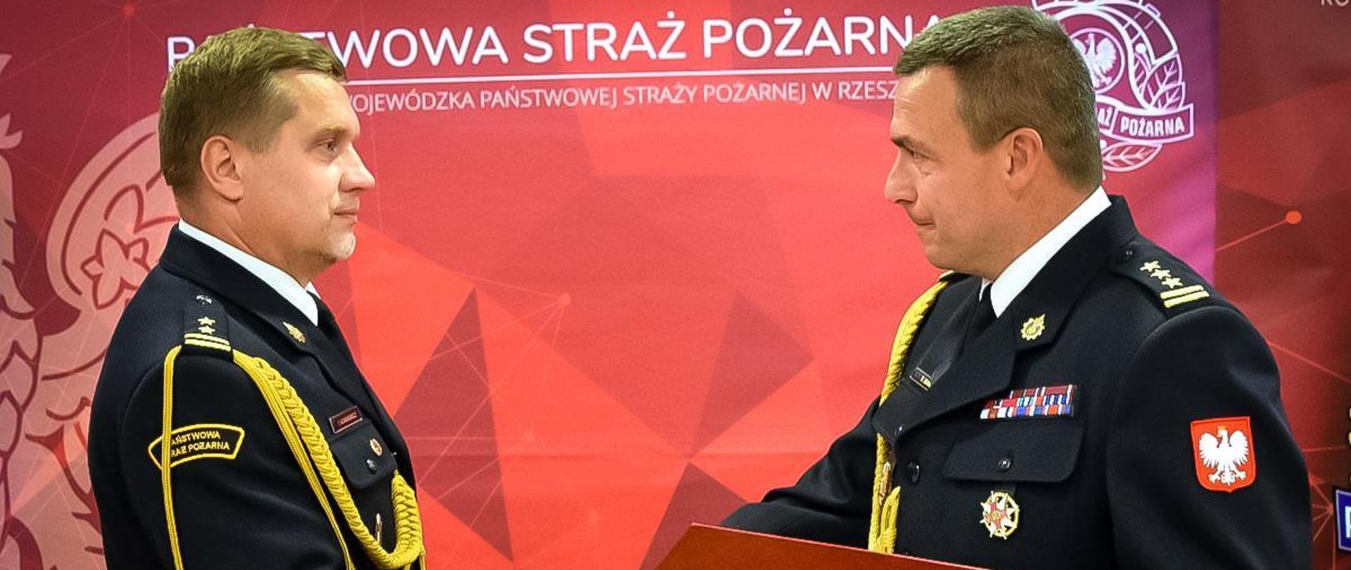 Zdjęcie przedstawia powołanie na stanowisko zastępcy komendanta miejskiego Państwowej Straży Pożarnej w Rzeszowie bryg. Tomasza Korniewicza. Akt powołania wręcza podkarpacki komendant wojewódzki PSP st. bryg. Tomasz Baran. Komendanci ubrani w mundury wyjściowe stoją naprzeciwko siebie z uściśniętymi dłońmi. W tle widoczny jest baner Państwowej straży Pożarnej.