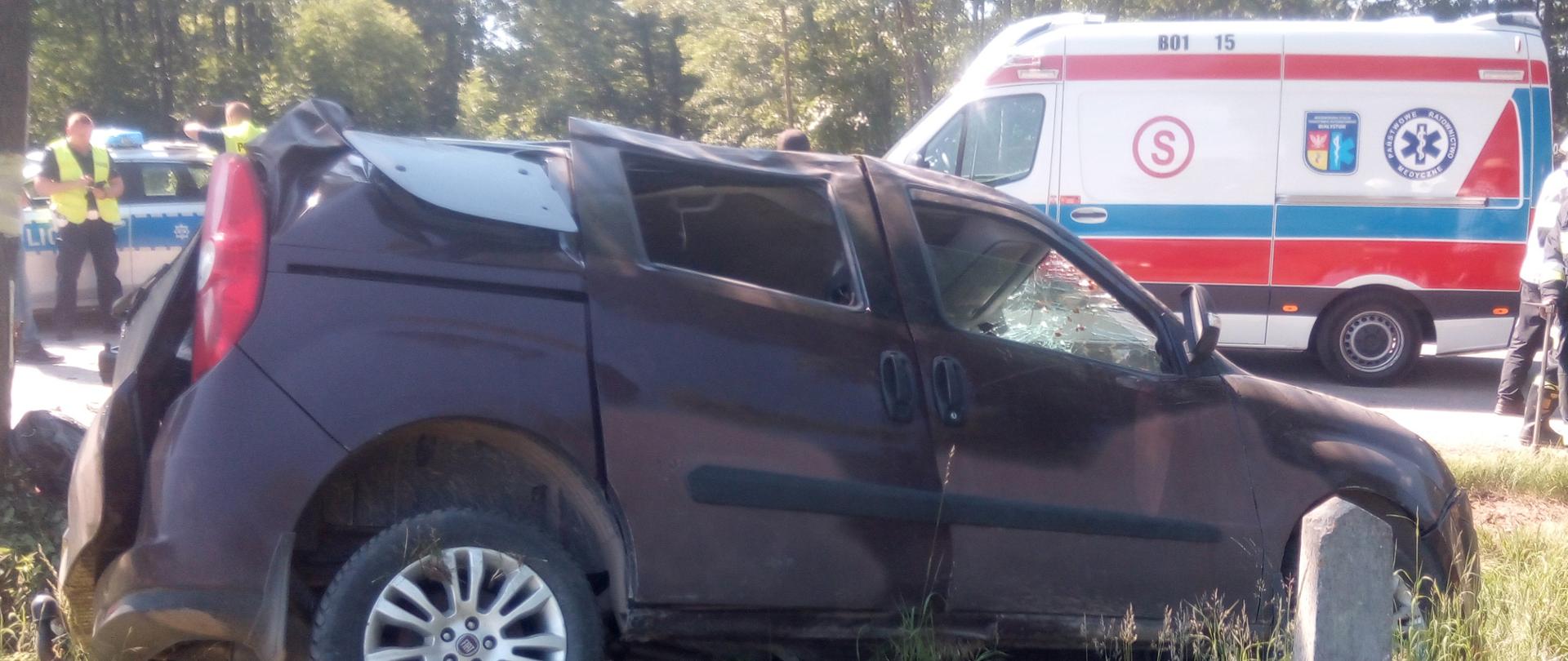 Uszkodzony samochód osobowy stojący przechylony na bok w przydrożnym rowie. W tle karetka oraz radiowóz.