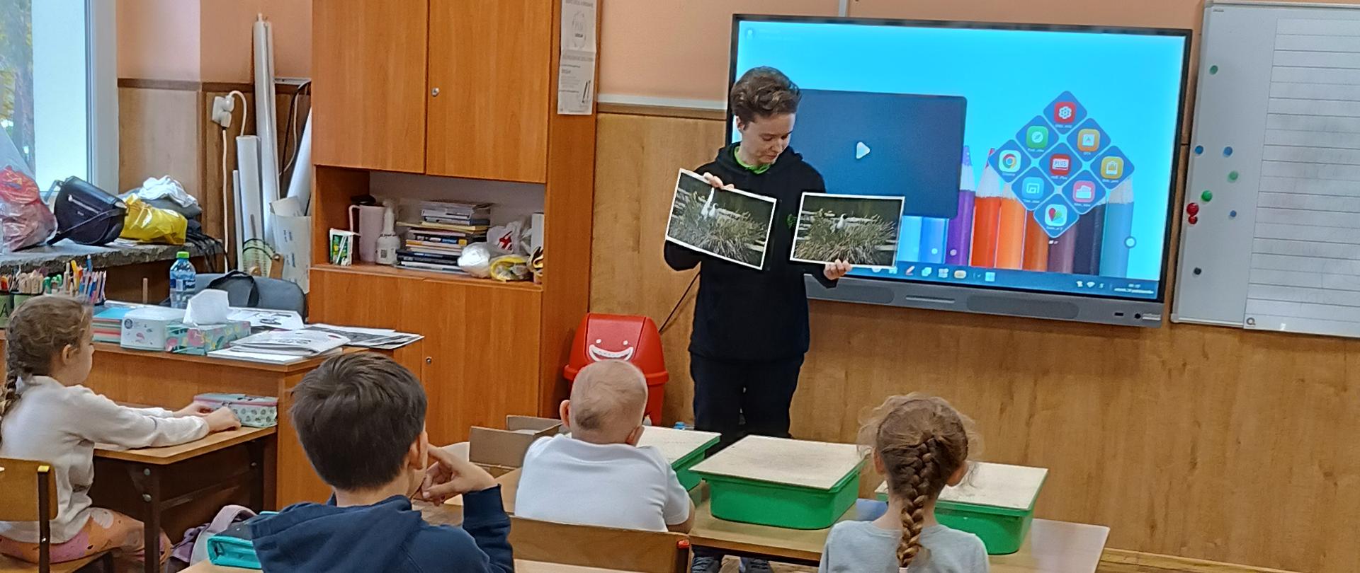 Na tle tablicy szkolnej stoi kobieta trzymająca dwa zdjęcia, tyłem przy ławkach siedzą dzieci