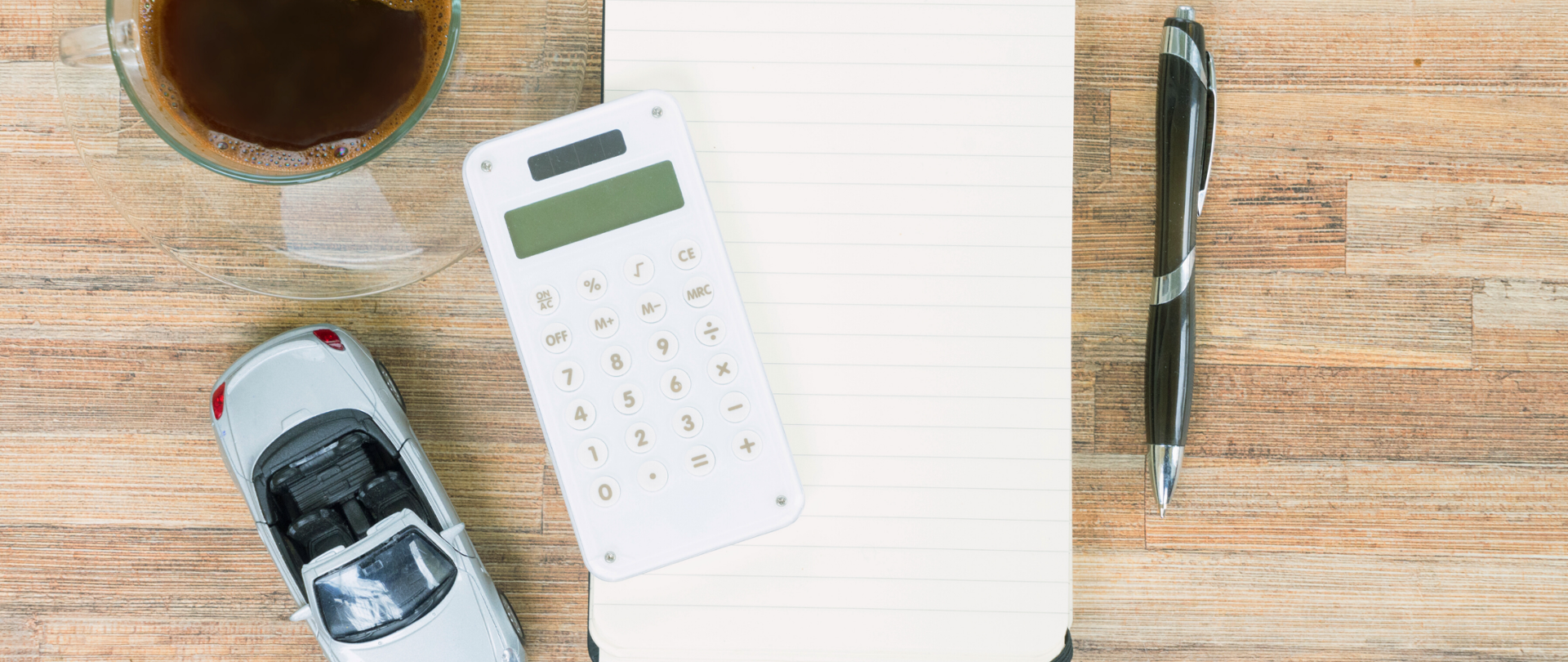 Biurko. Na środku leży notes w linie, na nim położony jest kalkulator. Po prawej stronie leży długopis, po lewej u góry szklanka z kawą, poniżej resorak - szary kabriolet.