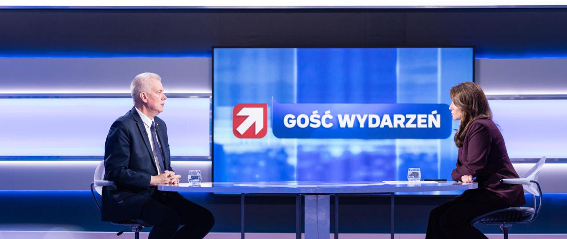 Na zdjęciu wnętrze studia telewizyjnego. Przy szklanym stole siedzą: minister Tomasz Siemoniak (po lewej) oraz redaktor Magda Sakowska (po prawej). Na stole szklanki z wodą. Pomiędzy osobami monitor - na niebieskim tle biały napis "GOŚĆ WYDARZEŃ" oraz biała strzałka na czerwonym tle. W tle zdjęcia poziome oświetlenie studia.