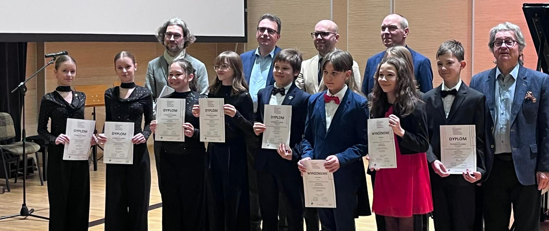 Zdjęcie grupowe laureatów XVII Ogólnopolskiego Konkursu Kameralistyki Fortepianowej w Toruniu. W rzędzie dalej stoją mężczyźni - jurorzy. Z przodu stoi 10 pianistów obu płci. Większość ubrana jest na czarno, wszyscy odświętnie. Jedna dziewczynka ma czerwoną spódnicę. Młodzież trzyma w ręku dyplomy A4.