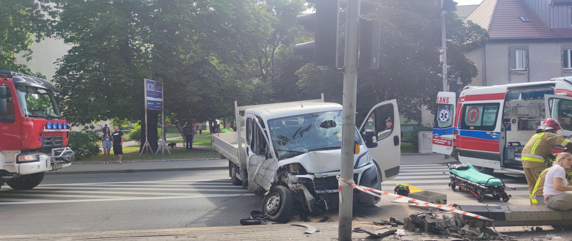 na zdjęciu widać rozbity samochód dostawczy. Po prawej stronie strażacy udzielają pomocy poszkodowanemu. Po lewej stronie widać przód samochodu strażackiego.