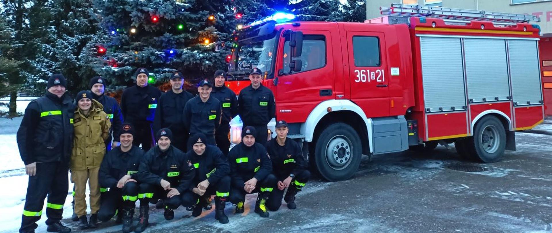 Grupa strażaków na tle choinki. Za nimi stoi wóz strażacki