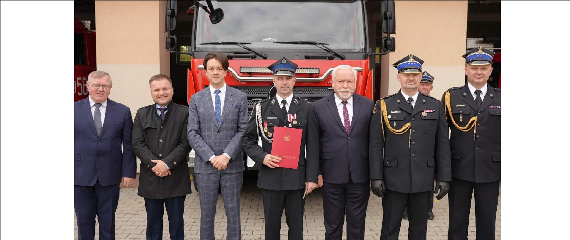 strażacy i zaproszeni gości stoją przed garażem OSP z aktem przekazania samochodu pożarniczego, w tle nowy samochód pożarniczy