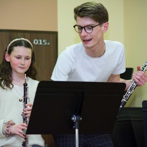 zdjęcie przedstawia prowadzącego warsztaty trzymającego w lewej dłoni obój, obok uczennica trzymająca obój, na pierwszym planie pulpit, prowadzący udziela wskazówek