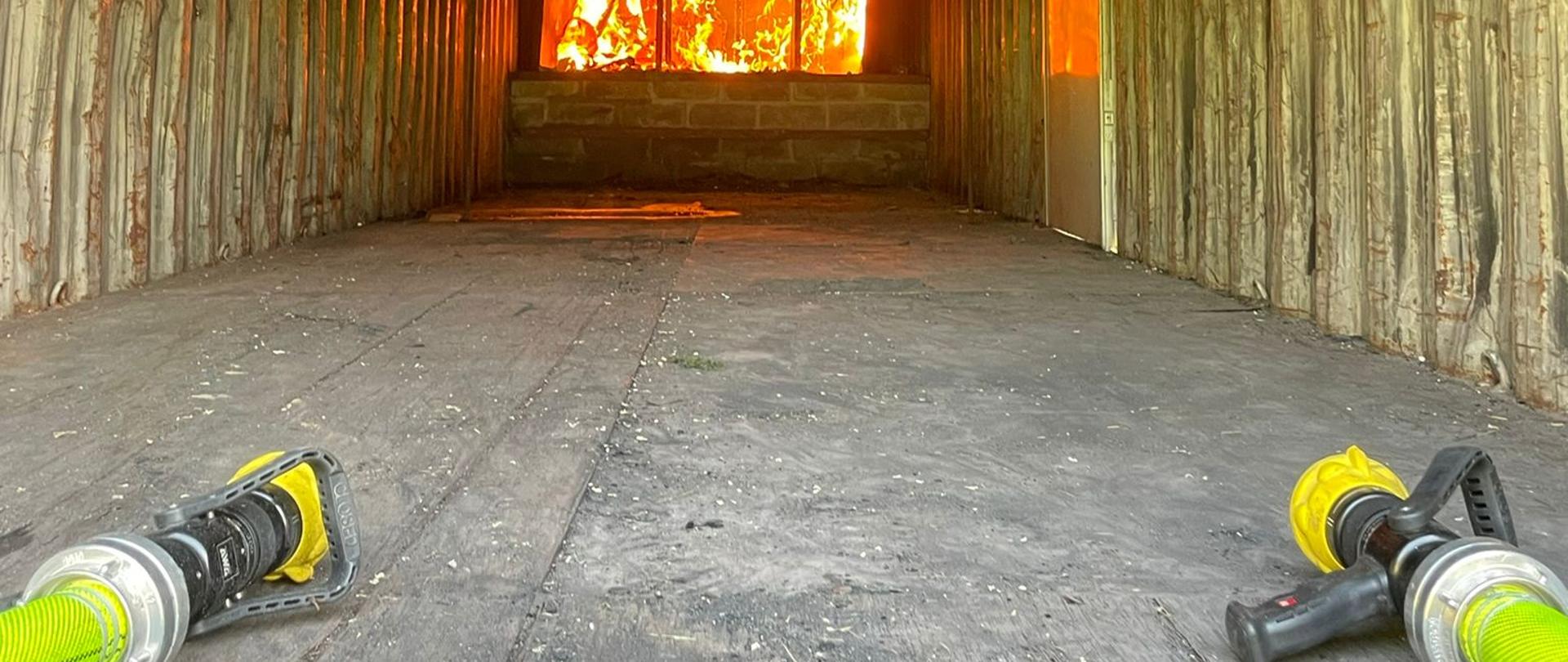 Na zdjęciu widzimy dwa węże gaśnicze w kontenerze pożarowym. Z oddali widać płomienie. Na ziemi miedzy nimi jest widoczna linia gaśnicza w kolorze żółtym. Przygotowanie do szkolenia z zakresu gaszenia pożarów wewnętrznych.
