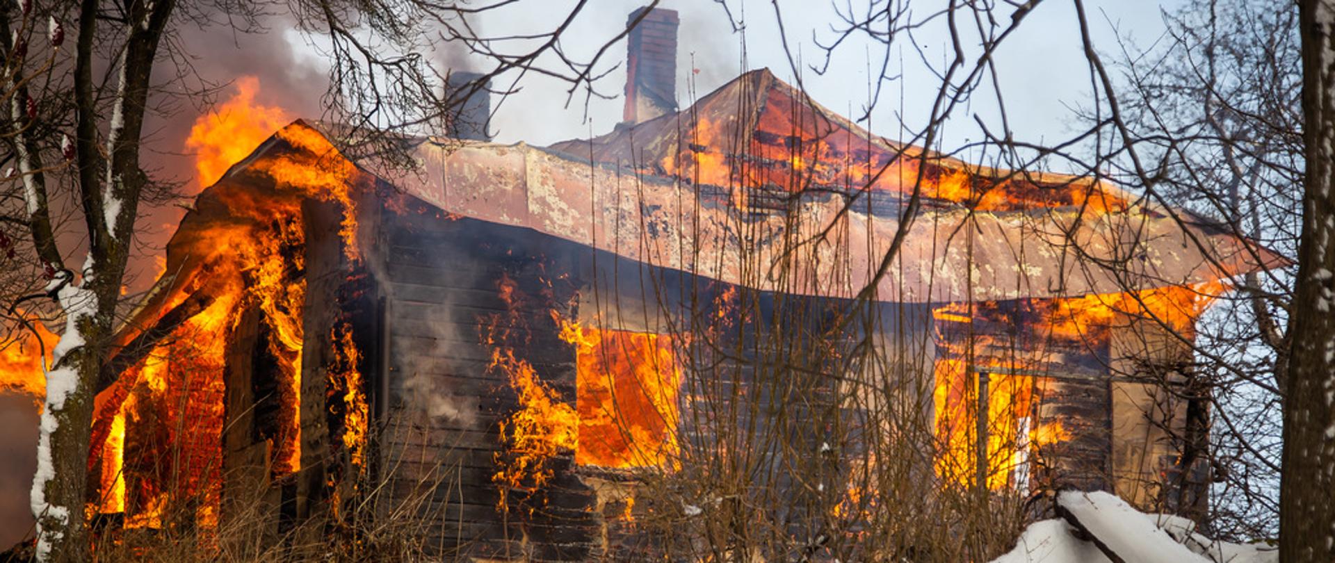Drewniany dom w całości objęty ogniem . Wokół dużo śniegu. Języki ognia wydostają się przez okna
