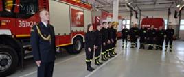 Na garażu obok pożarniczego samochodu bojowego w rzędzie stoją strażacy ubrani w mundury koszarowe. W tle inni strażacy również w mundurach koszarowych