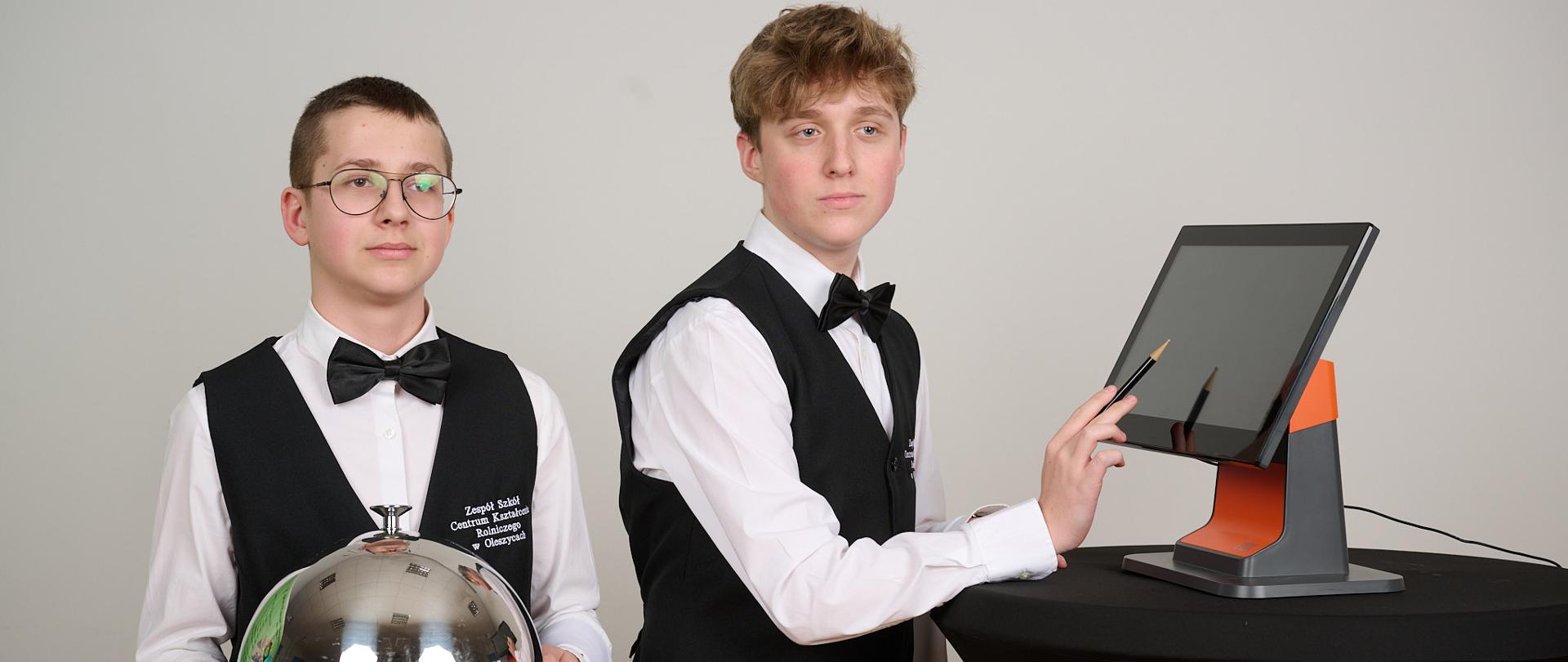 Dwóch chłopców stoi w strojach kelnerskich, patrzą w lewym kierunku. Jeden trzyma tacę srebrną w lewej ręce, a na drugiej ma przewieszoną serwetę. Drugi trzyma dłoń przy monitorze, wspartą na stoliku, a w dłoni ołówek.