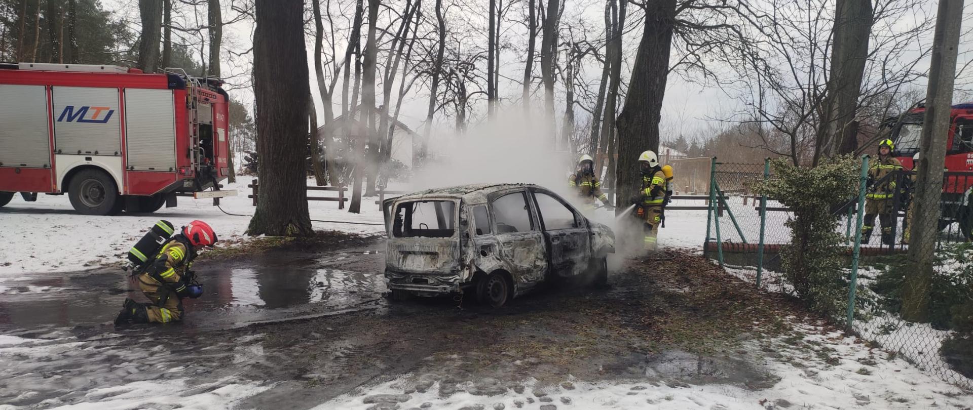 Całkowicie spalony samochód osobowy, strażacy dogaszający samochód, w tle samochody pożarnicze